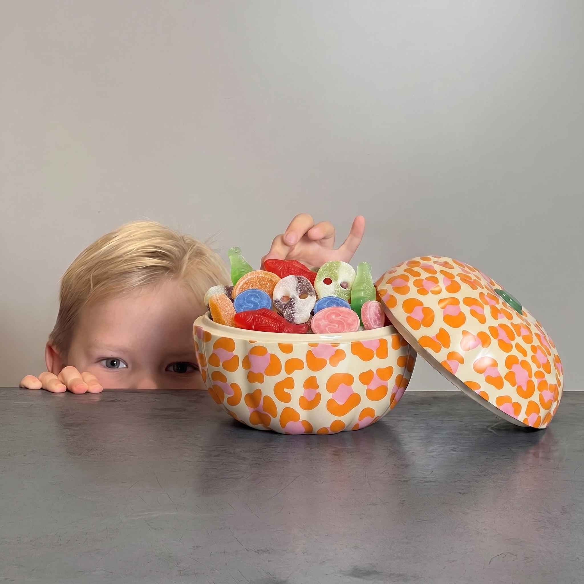 A child's face peeks over a table at a pumpkin shaped tin with a matching lid, both decorated in an orange and pink leopard print. The tin is filled with colorful swedish candies, set against a plain gray background.