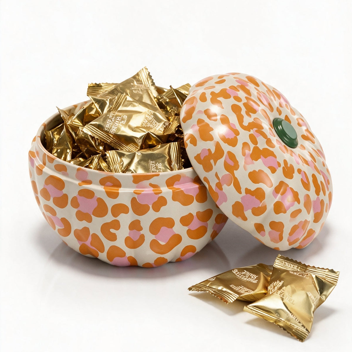 A pumpkin shaped tin with a lid, decorated in a pink and orange leopard print pattern on a cream background. The tin is filled with gold foil wrapped chocolate truffles, with two loose truffles in the foreground on a clean white background